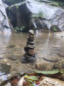 A small stack of balanced stones in a shallow, clear stream with smooth, moss-covered rocks and some vegetation in the background. Leaves and pebbles are visible under the water.
