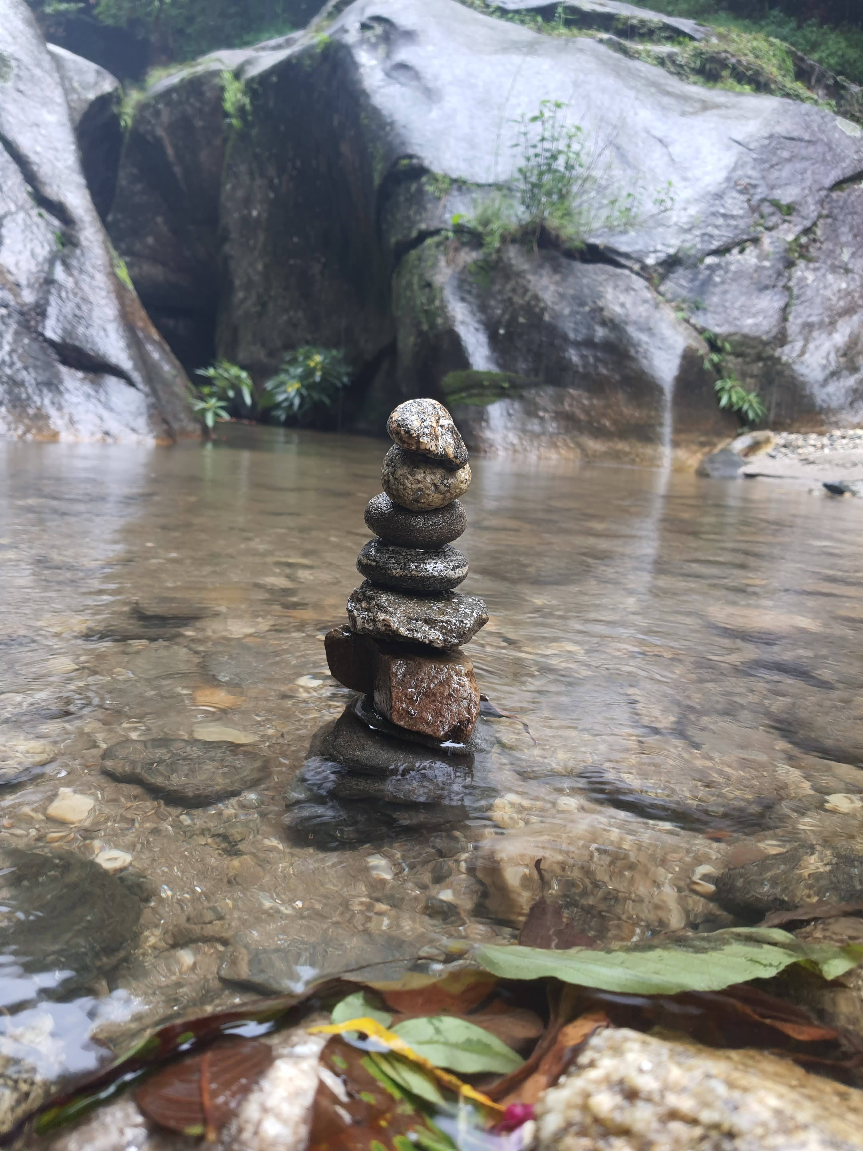 A small stack of balanced stones in a shallow, clear stream with smooth, moss-covered rocks and some vegetation in the background. Leaves and pebbles are visible under the water.