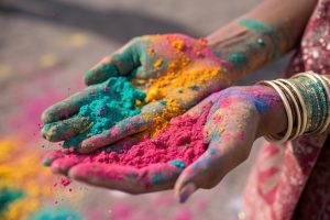 Two hands are held open, displaying vibrant colored powders in shades of turquoise, pink, and yellow. The hands are adorned with traditional bangles, and the skin is stained with remnants of the powders, suggesting a festive atmosphere. In the background, splashes of the same colors can be seen scattered on the ground, indicative of a celebration, 