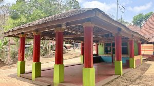 A small open-air structure with a tiled roof supported by red pillars with green bases, set against a backdrop of trees and a clear sky. The building has colorful walls in green, red, and blue, with decorative elements and a barred window. The surrounding area includes a mix of paved and unpaved ground, with other traditional buildings visible in t