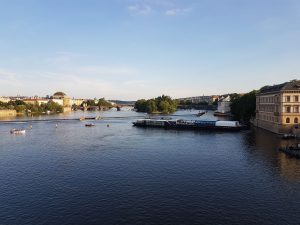 A scenic view of a river with boats and a bridge in the background, flanked by historic buildings and greenery under a clear blue sky.