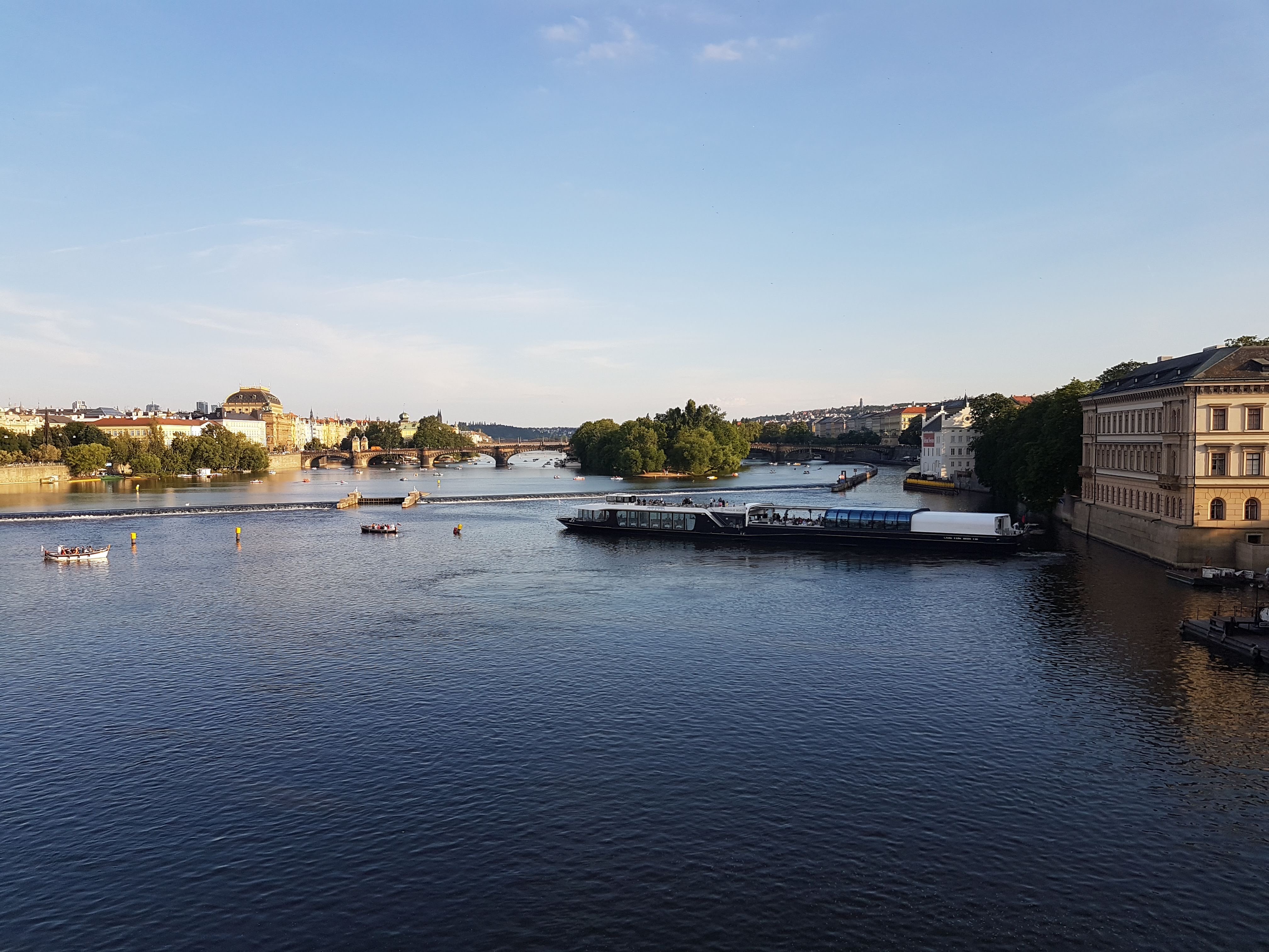 A scenic view of a river with boats and a bridge in the background, flanked by historic buildings and greenery under a clear blue sky.