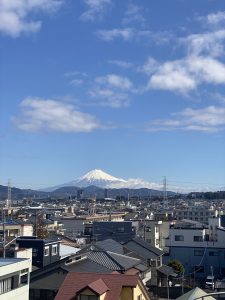 A panoramic view of a cityscape featuring a mix of residential buildings with varying architectural styles, set against a backdrop of a clear blue sky. In the distance, a snow-capped Mount Fuji dominates the horizon, lending a serene and majestic quality to the scene. The foreground includes rooftops and utility poles, adding depth to the urban landscape.