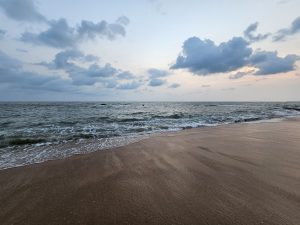 Kappad Beach in Kozhikode, Kerala, under a cloudy sky at dusk. The waves gently roll onto the sandy shore, creating a tranquil beach scene.


