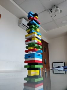 A colorful tower of wooden blocks stacked on a reflective glass table with a room’s ceiling and a projector visible above.
