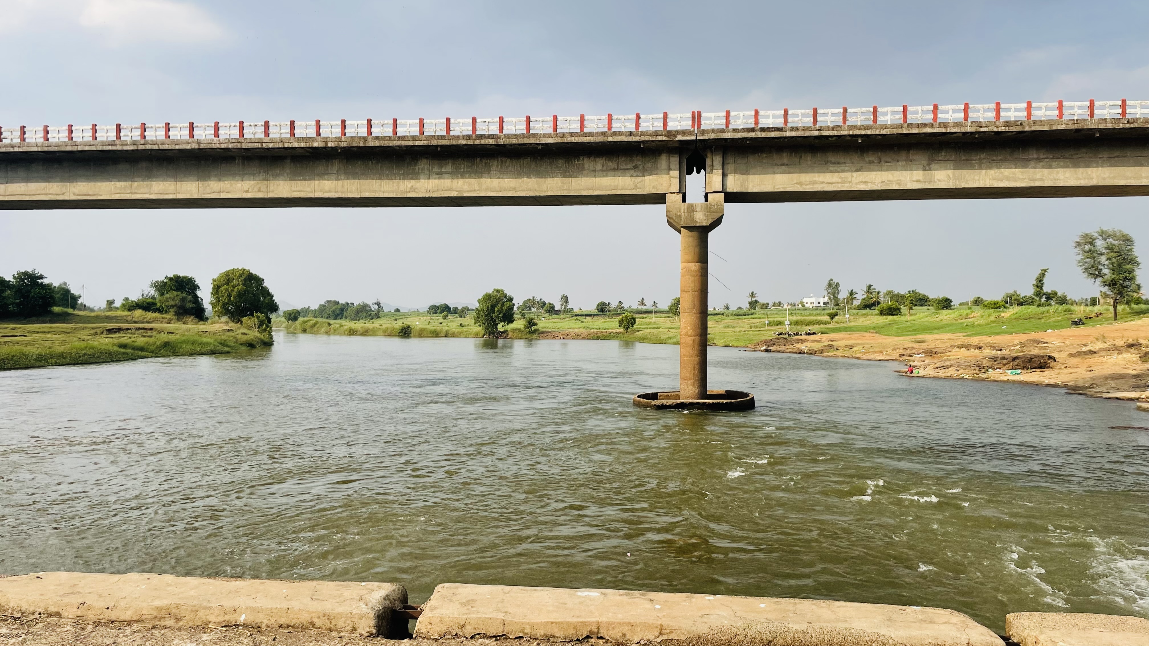 A concrete bridge over a flowing river with grassy banks and a cloudy sky.