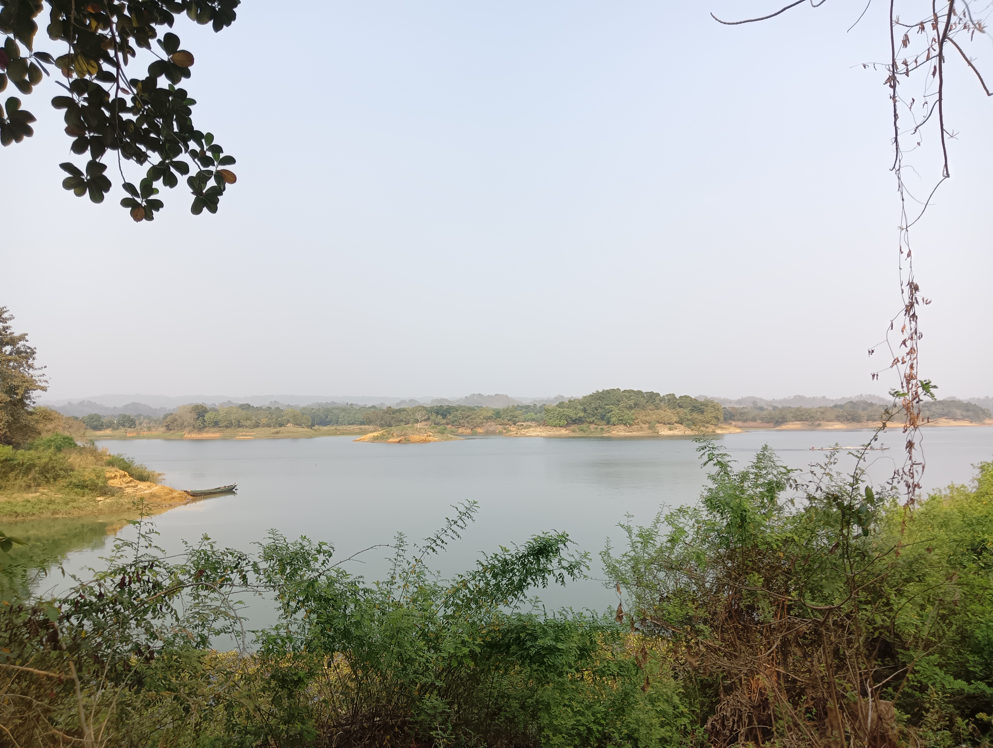 A tranquil landscape featuring a calm body of water surrounded by lush greenery and distant hills under a clear sky. In the foreground, there are various shrubs and plants, while a small boat is moored near the shoreline on the left side.