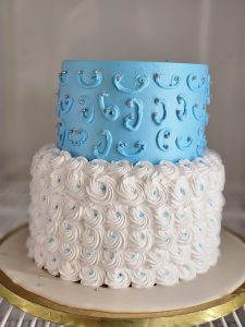 A two-tiered cake, with the top tier frosted in light blue with silver and blue accents, and the bottom tier covered in white rosettes with similar accents. The cake sits on a gold-trimmed base and has a soft-focus background.
