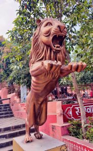 A bronze lion statue with its mouth open and front paws raised, set on a pedestal outdoors with green foliage and red steps in the background.