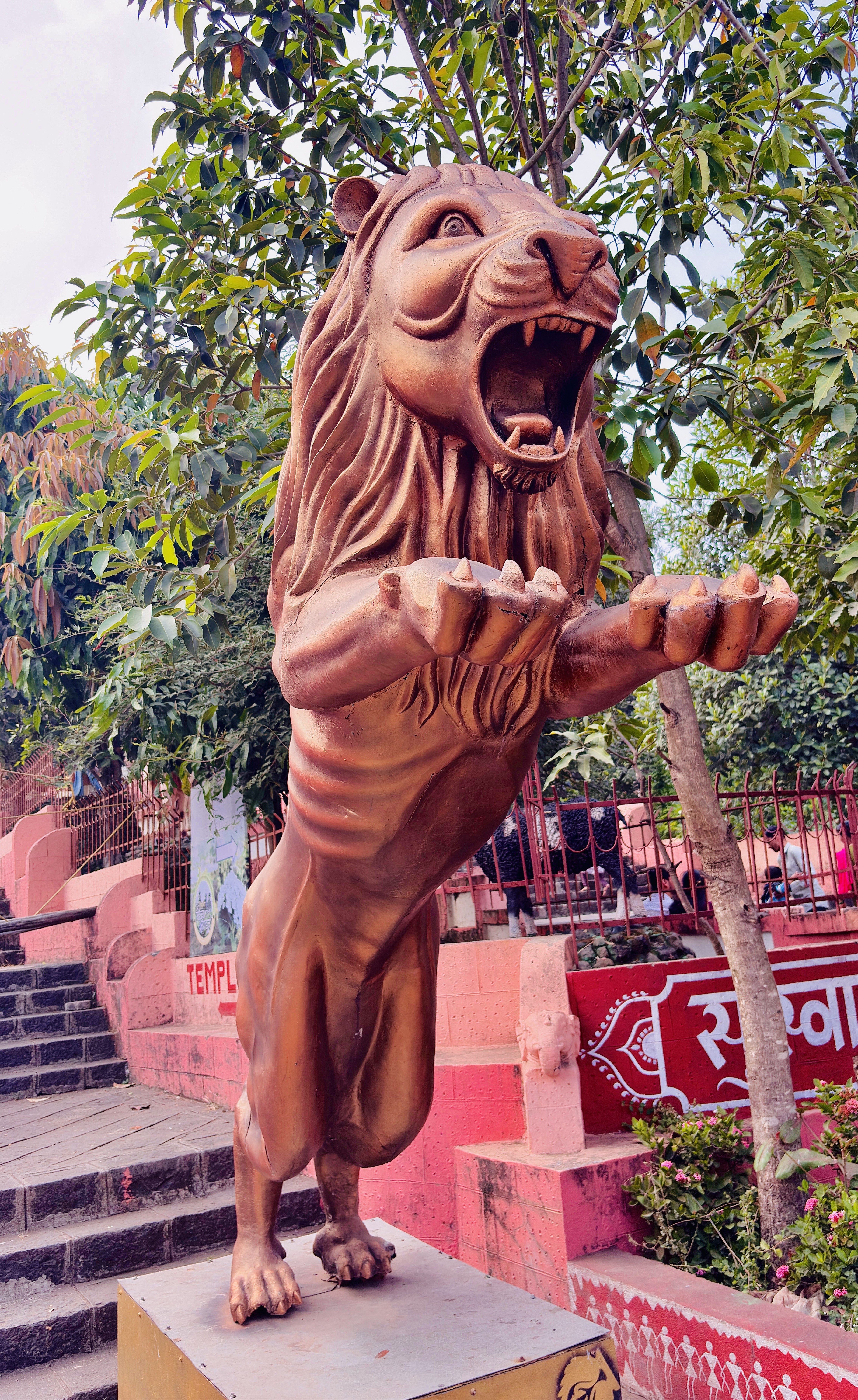 A bronze lion statue with its mouth open and front paws raised, set on a pedestal outdoors with green foliage and red steps in the background.