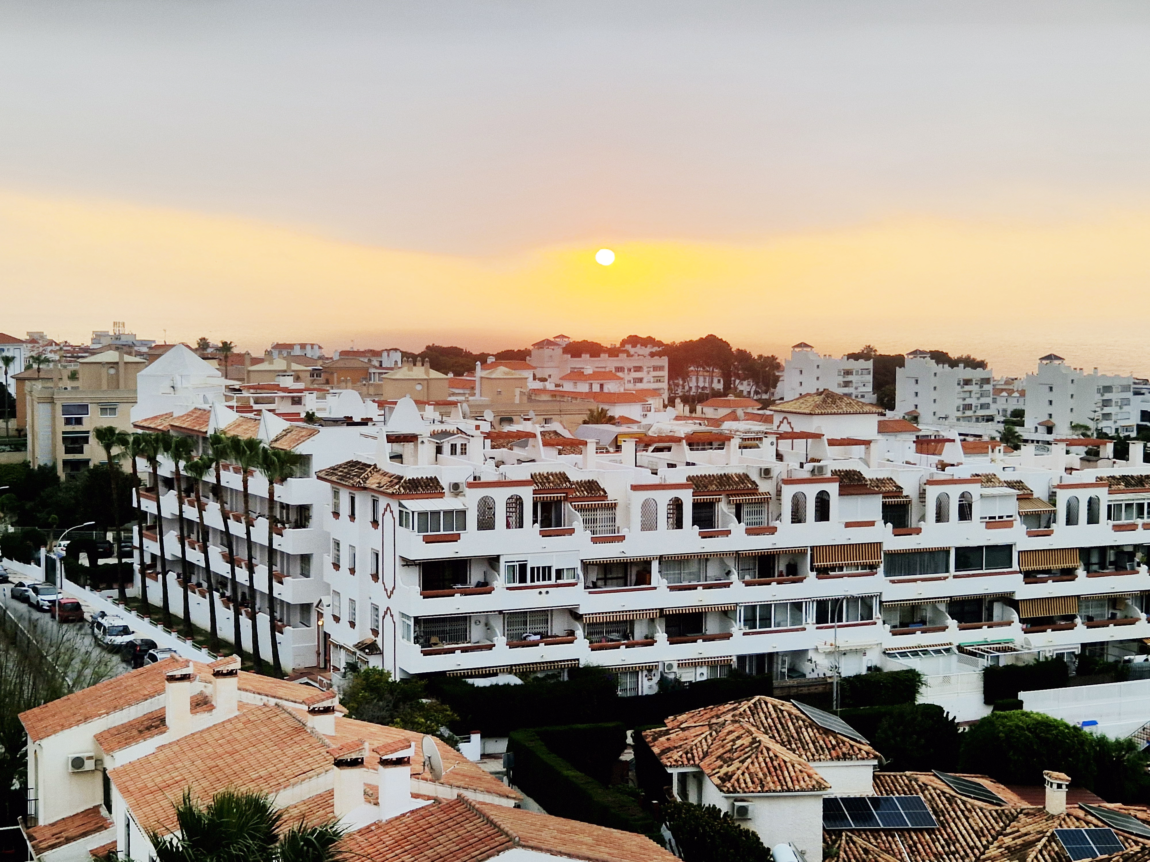 

A scenic view of a coastal town during sunrise, featuring a skyline of predominantly white buildings with terracotta roofs. Palm trees line the streets, and the sun sets over the horizon, casting a warm glow in the sky.