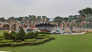 A large outdoor sculpture garden depicting a scene with multiple colorful statues of people, including a central figure seated on a multi-headed serpent. The background features intricately sculpted trees and rocks, creating a natural setting.