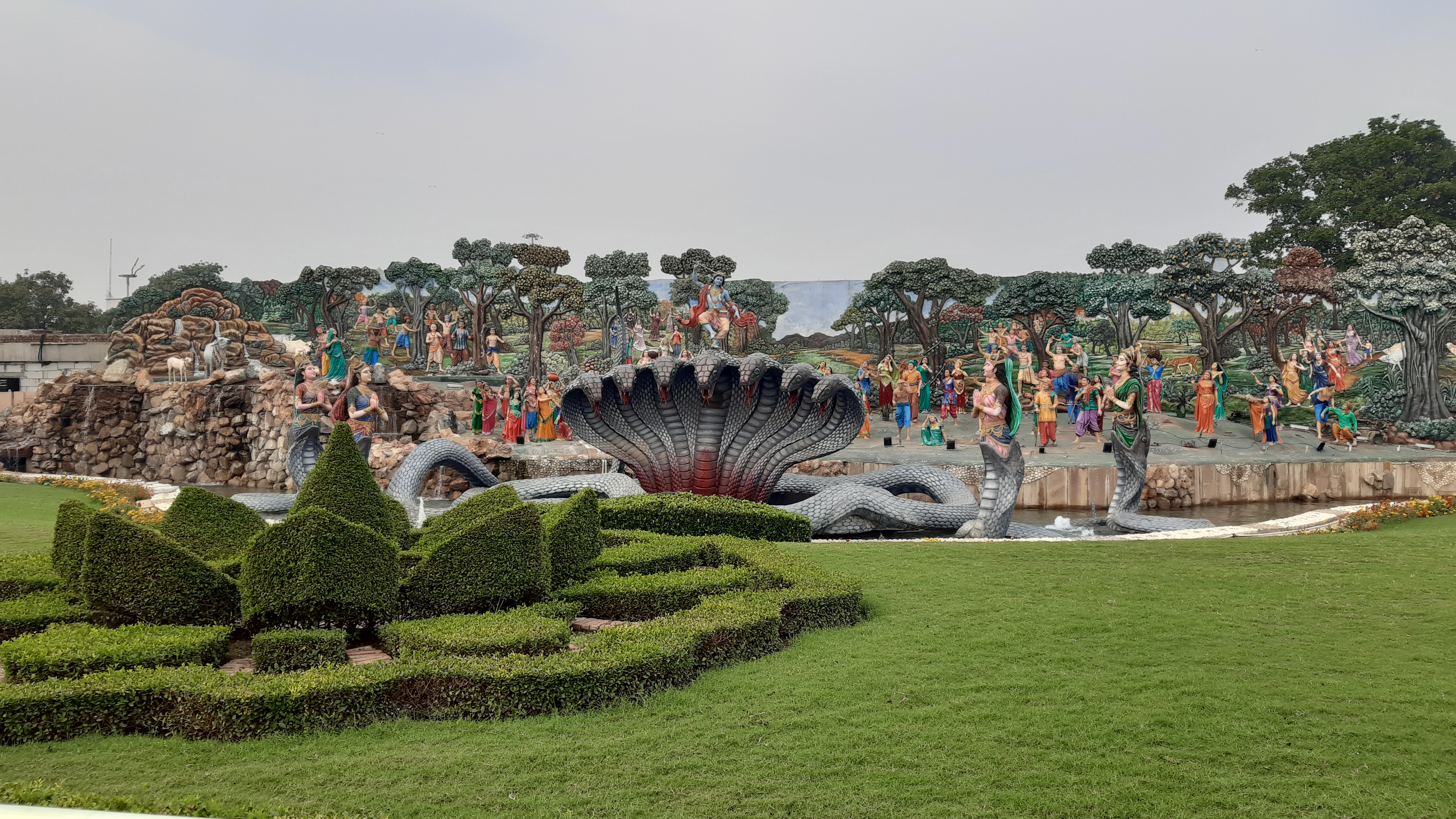 A large outdoor sculpture garden depicting a scene with multiple colorful statues of people, including a central figure seated on a multi-headed serpent. The background features intricately sculpted trees and rocks, creating a natural setting.