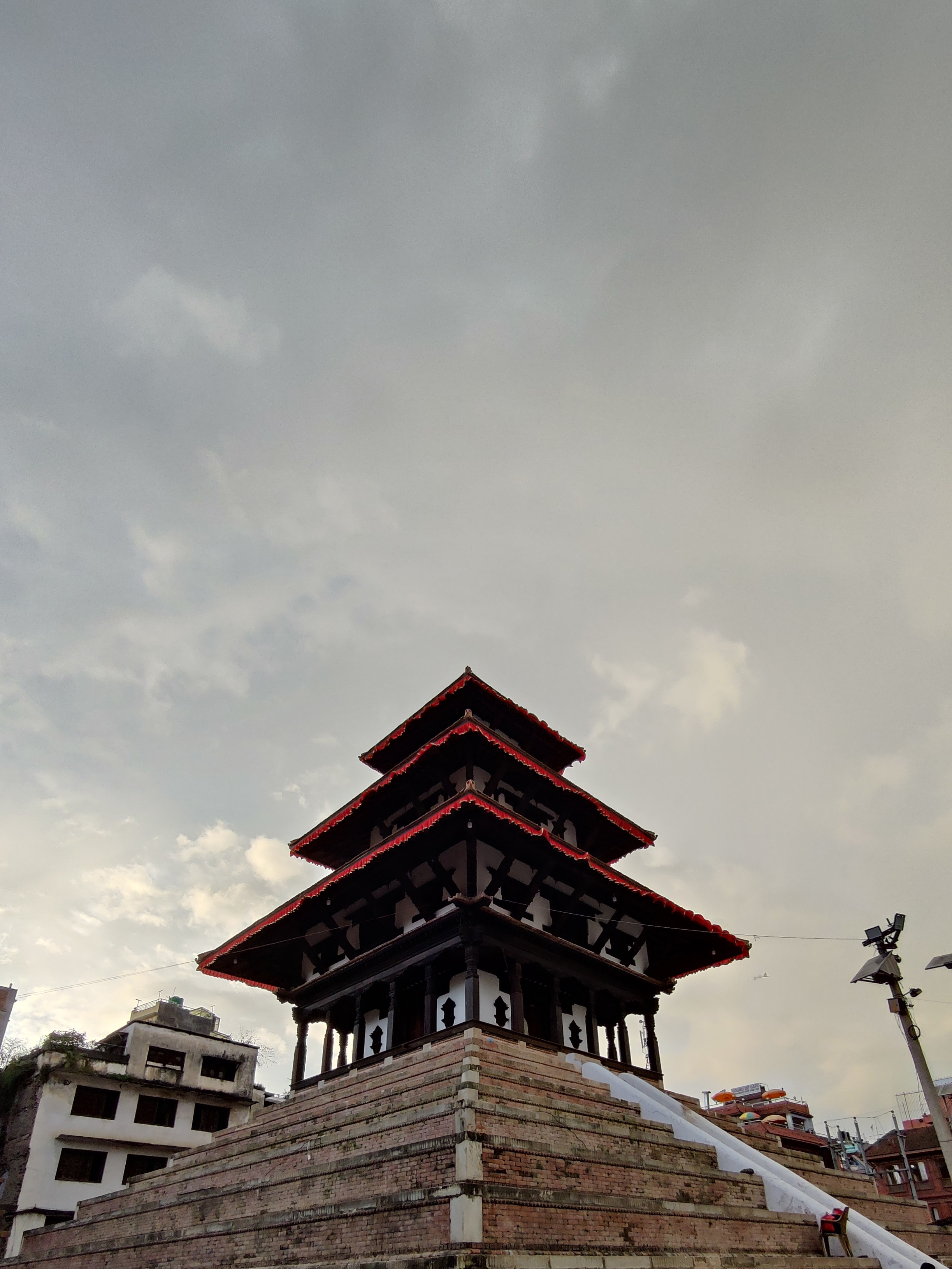 A traditional multi-tiered pagoda-style temple with red and black accents stands atop a large brick platform against a cloudy sky. Surrounding buildings and a lamppost are visible in the background.