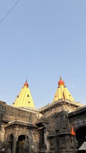 Ancient stone temple structure with two tall, yellow pyramid-shaped towers topped with red domes and golden spires, set against a clear blue sky. The foreground shows intricately carved dark stone walls and sculptures, along with an orange flag mounted on a pole.