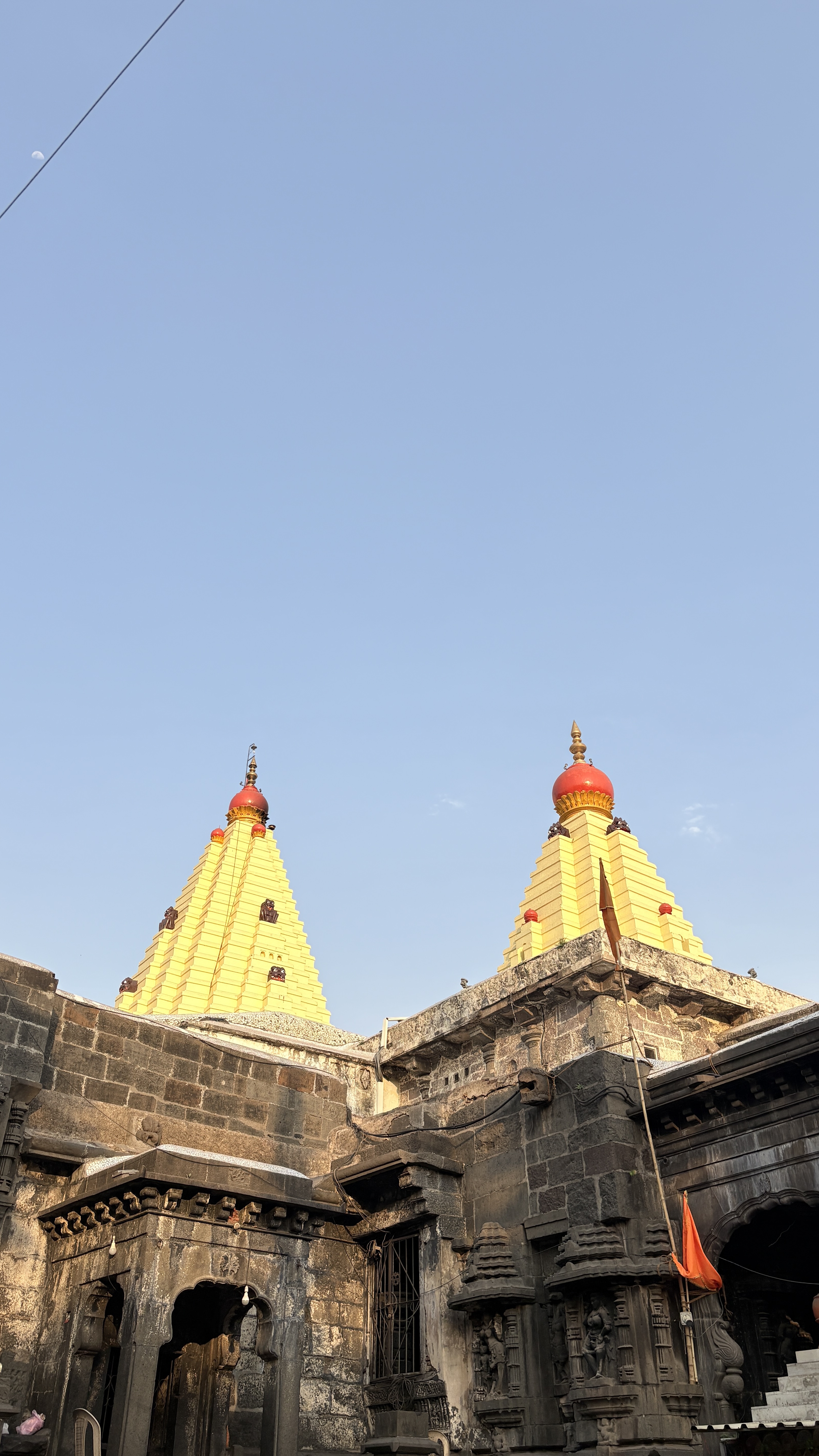 Ancient stone temple structure with two tall, yellow pyramid-shaped towers topped with red domes and golden spires, set against a clear blue sky. The foreground shows intricately carved dark stone walls and sculptures, along with an orange flag mounted on a pole.