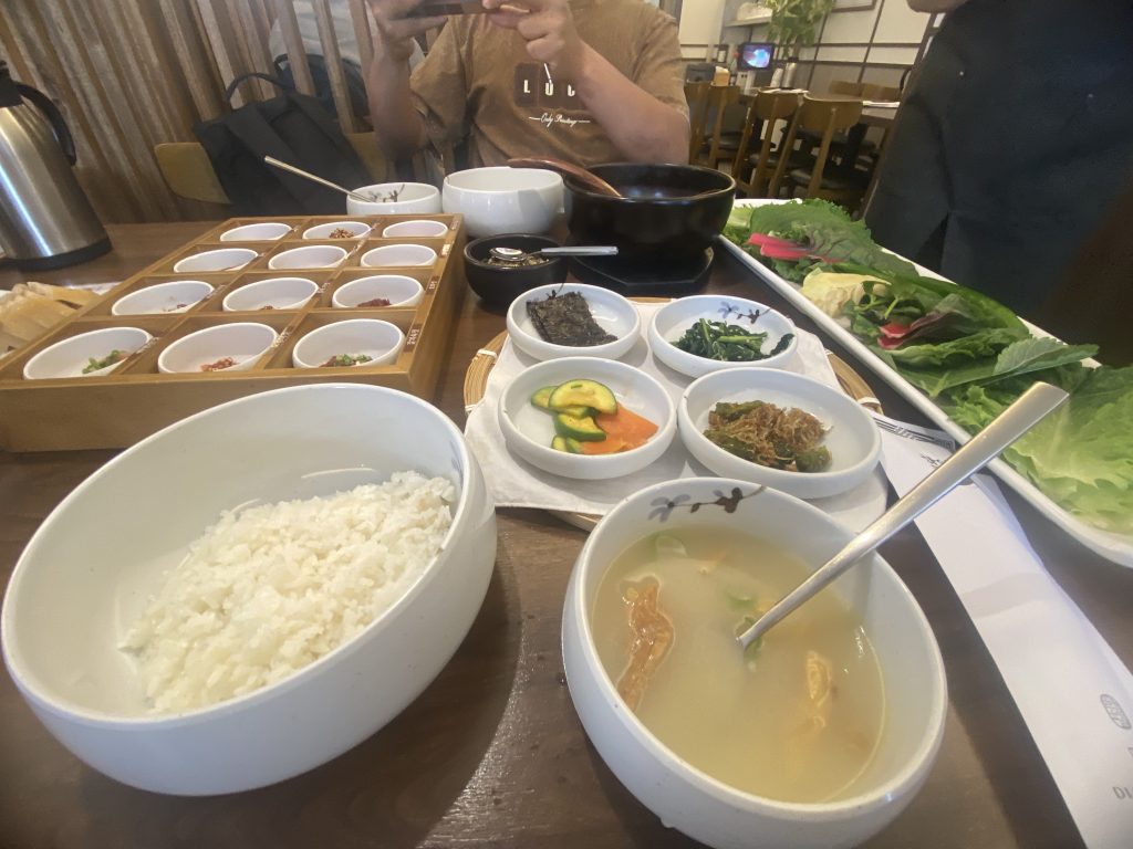 A table setting featuring a variety of dishes: a bowl of white rice, a bowl of light soup with green onions and shredded ingredients, and several small bowls containing side dishes including vegetables and sauces. In the background, there is a wooden tray with additional small bowls, likely for condiments.