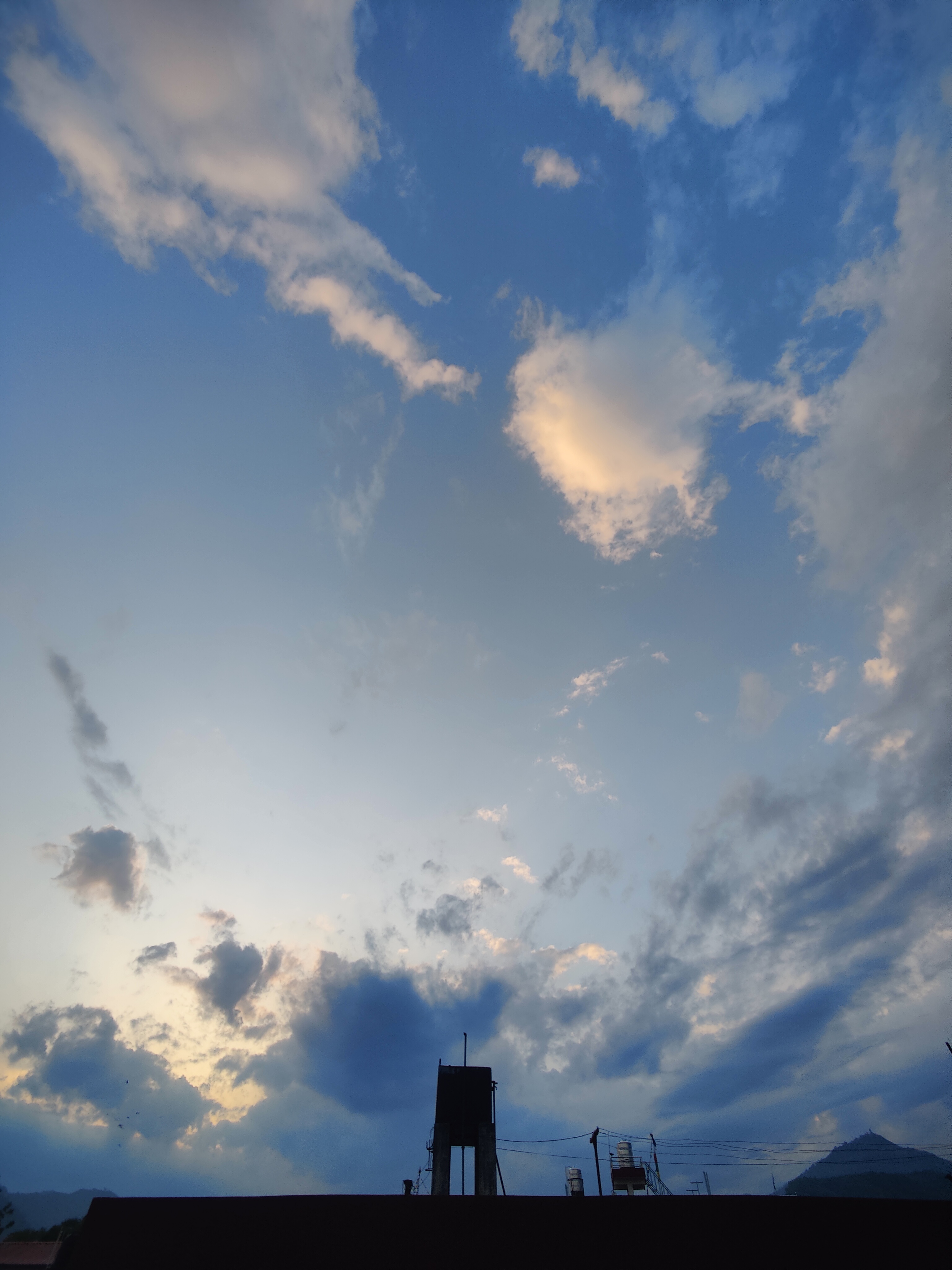 A sky during sunset, featuring scattered clouds illuminated by soft evening light. Below is a silhouette of a structure with wires and a distant hill visible, creating a serene and atmospheric scene.