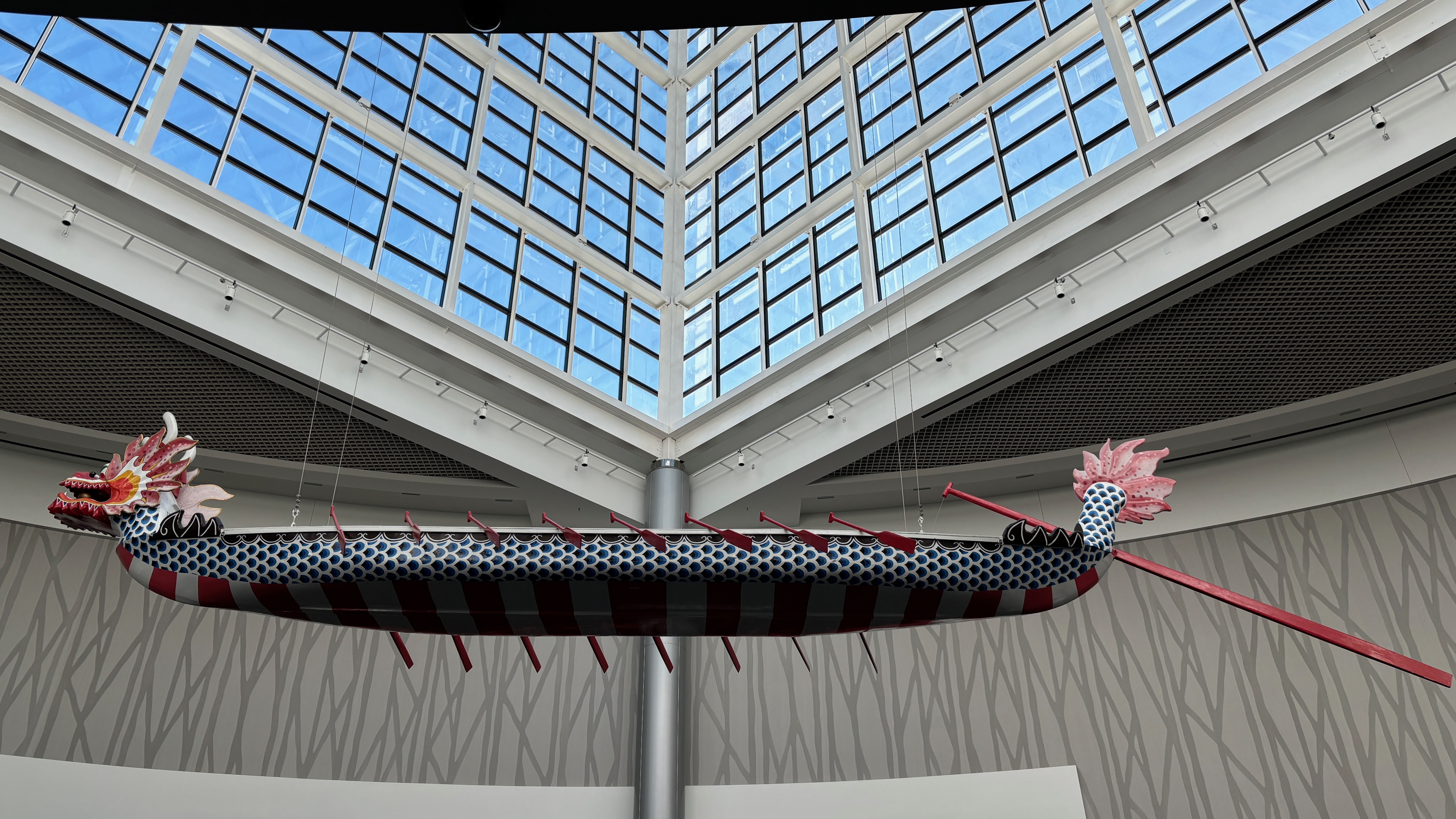 A long, decorative dragon boat is suspended from the ceiling inside a large glass atrium. The boat features a colorful dragon head and tail, with a pattern of blue scales and red and white stripes along its body. Red paddles are attached to its sides, and the background shows the structural beams and windows of the atrium.