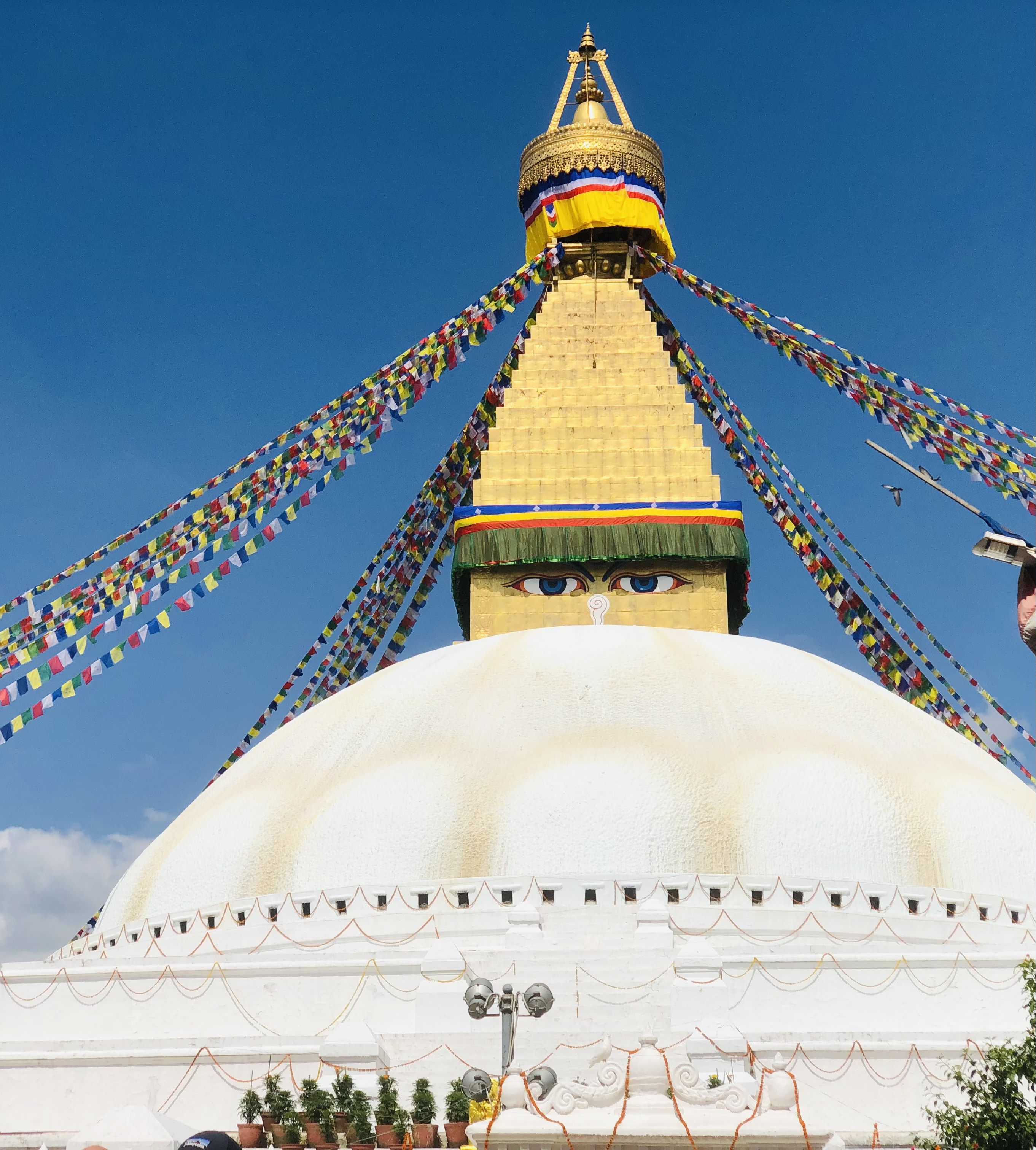 A large white stupa with a gilded spire and colorful prayer flags strung from the top. The stupa features the iconic Buddha eyes and a blue sky in the background.