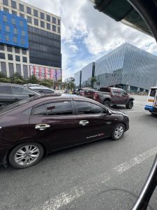 A city street scene featuring a dark-colored sedan and other vehicles in traffic, with modern office buildings and a cloudy sky in the background.