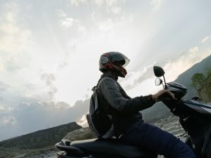 A person wearing a helmet and backpack is riding a scooter on a rocky terrain, with a hilly landscape in the background.