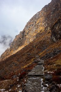 A stone pathway ascends through a mountainous landscape with rocky terrain and sparse vegetation, under an overcast sky with some clouds.
