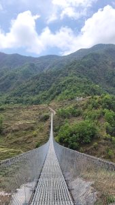 A long suspension bridge with a metal walkway stretches across a valley, leading towards a mountainous landscape covered in dense green trees. The sky above is partly cloudy, with patches of blue peeking through. In the background, the mountains rise steeply, and a small white building is visible on a hillside. The bridge appears to be in a remote and natural setting, inviting exploration into the surrounding scenery.