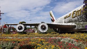 A large airplane structure covered in white and yellow flowers with the letters "Emi" visible on the side, set amidst a vibrant garden with colorful flowers under a partly cloudy sky.
