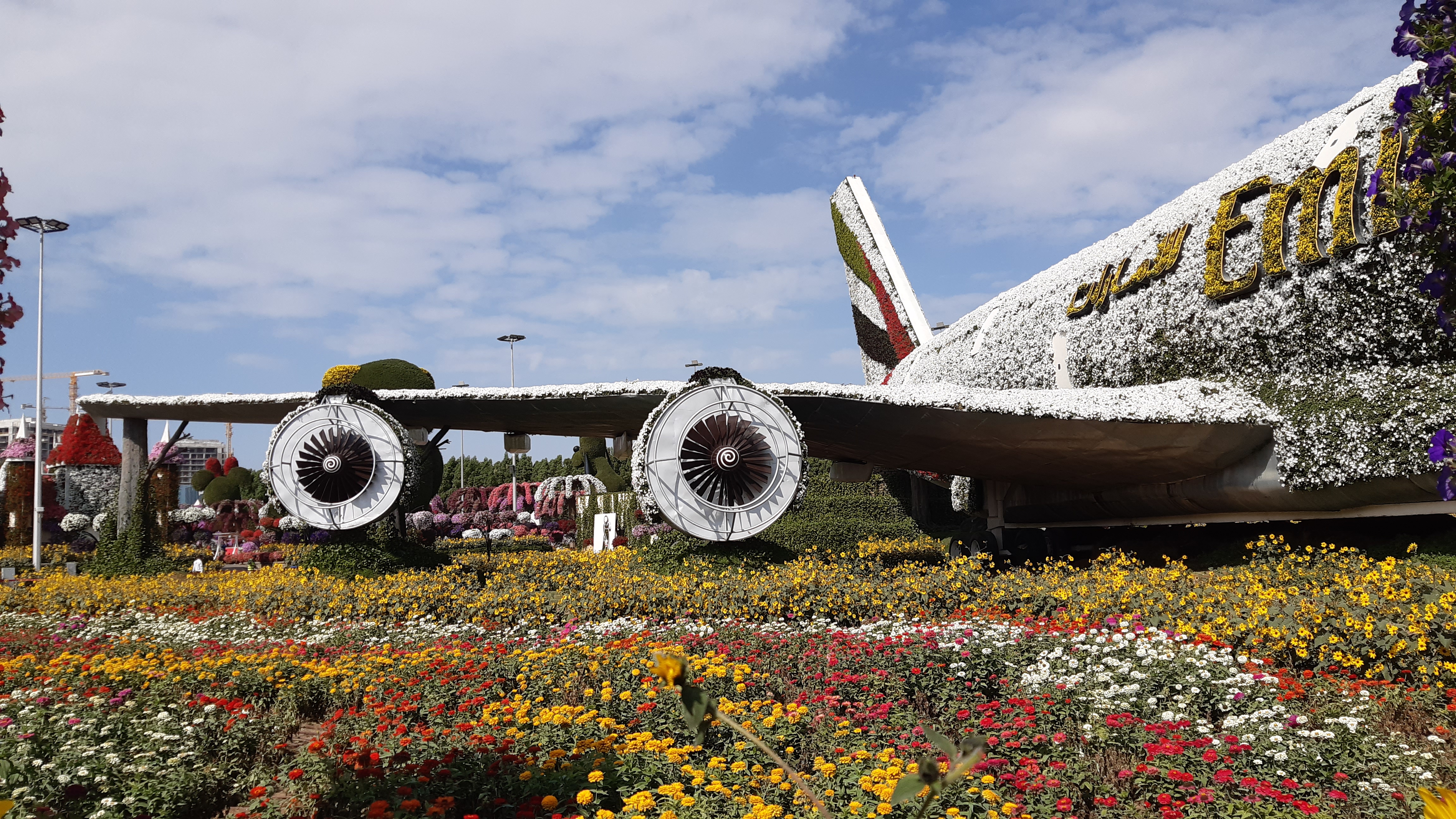 A large airplane structure covered in white and yellow flowers with the letters "Emi" visible on the side, set amidst a vibrant garden with colorful flowers under a partly cloudy sky.