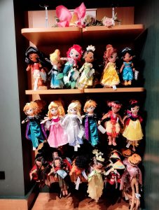 A display of colorful plush dolls, each dressed in various themed costumes, arranged on three wooden shelves. The top shelf holds six dolls, the middle shelf seven, and the bottom shelf has more dolls hanging by their arms.