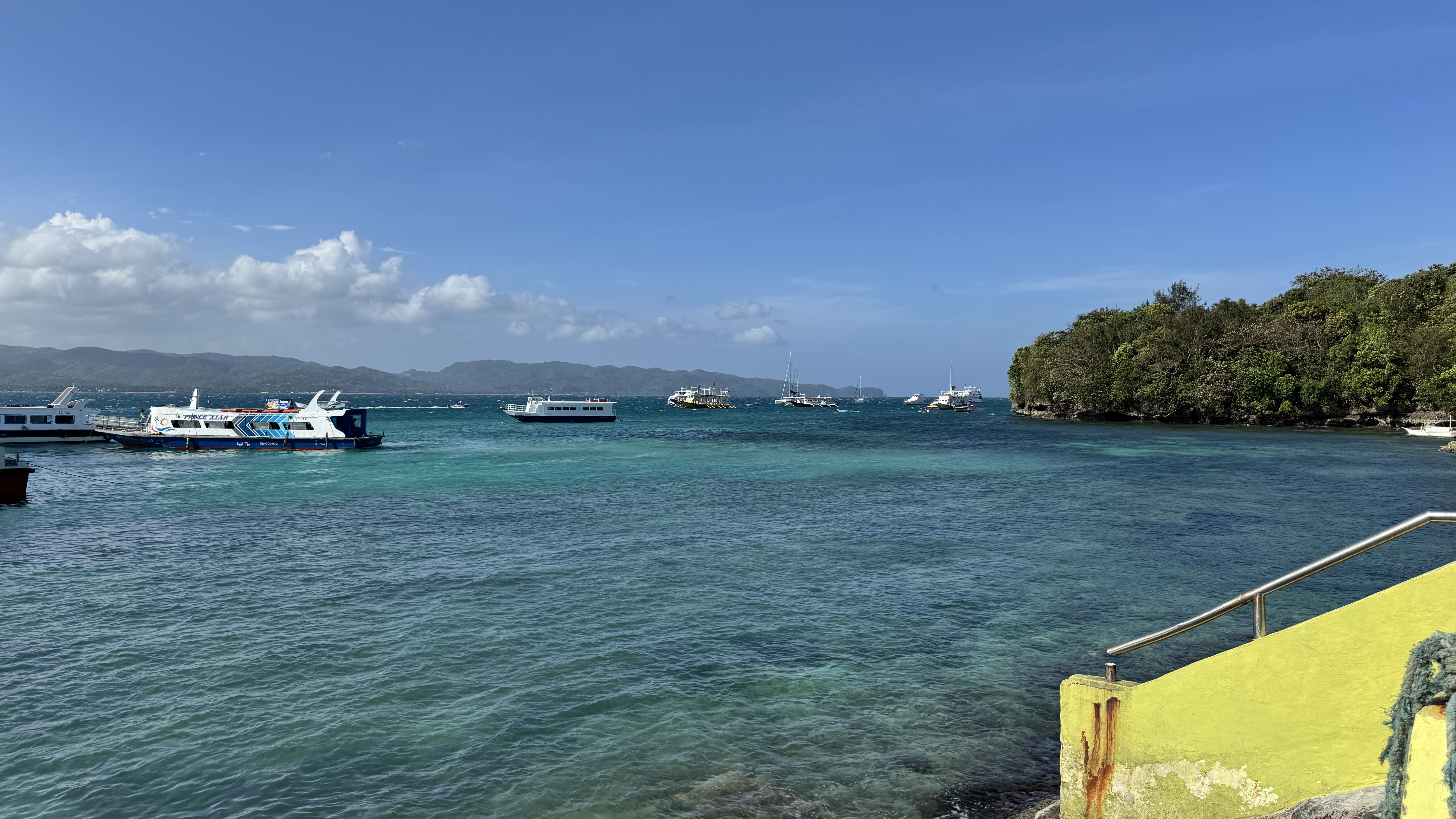 A scenic view of a coastal area with clear turquoise water, boats floating on the sea, and a lush green shoreline in the background under a clear blue sky