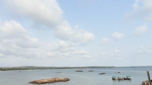A serene coastal scene with a calm sea under a partly cloudy sky. Several small boats with green canopies are anchored near the shore, while rocky outcrops jut out from the water. In the distance, a sandy beach stretches along the coastline, backed by lush greenery and rolling hills.
