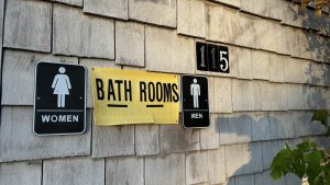 A yellow sign that says Bath Rooms between signs that say Men and Women next to a placard that say 115 on a wall made of wood shakes.