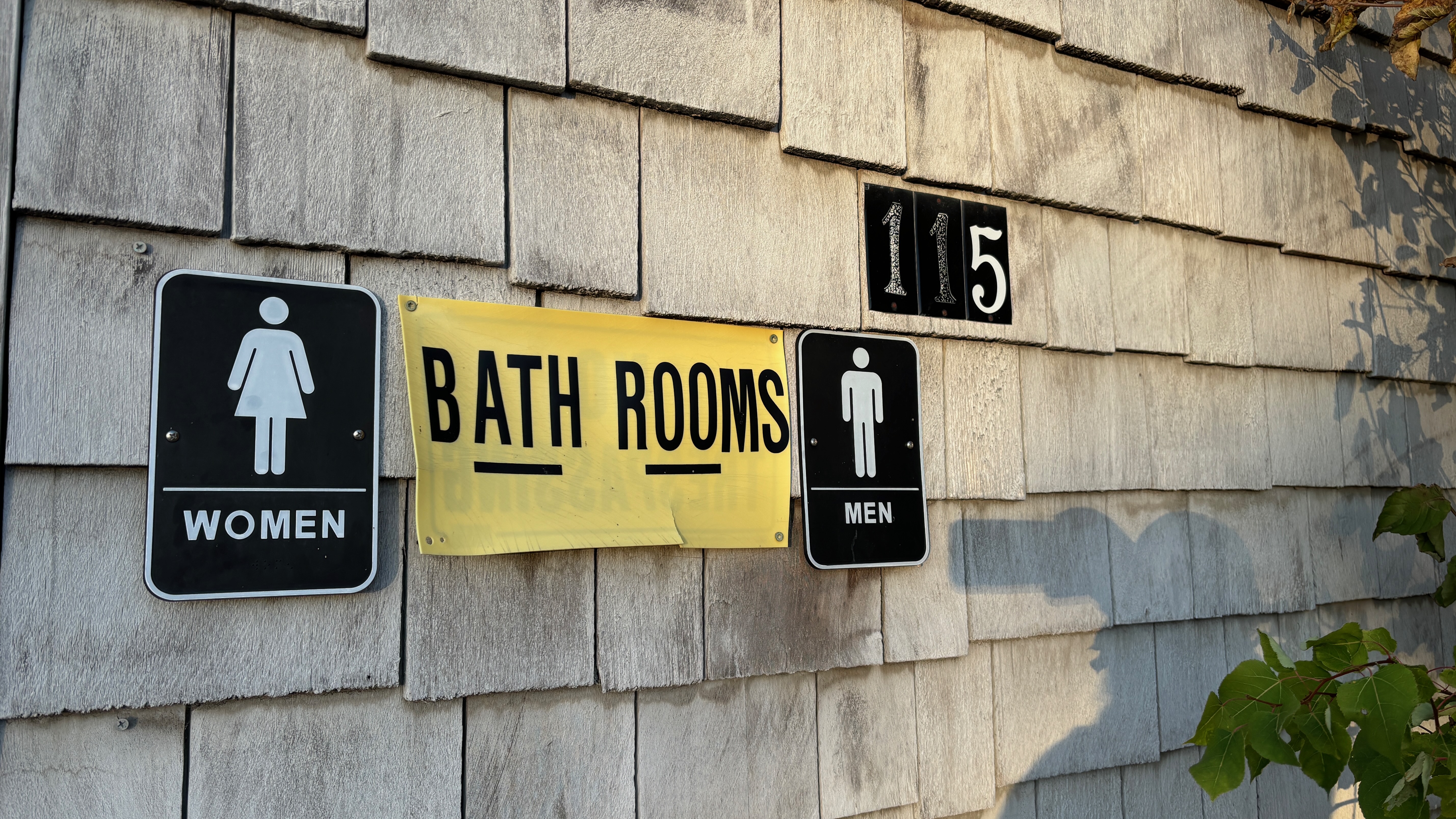 A yellow sign that says Bath Rooms between signs that say Men and Women next to a placard that say 115 on a wall made of wood shakes.