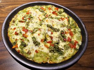 A freshly baked Pesto Chicken pizza on a dark gray plate, featuring a green pesto base, melted cheese, and toppings of red bell peppers and asparagus. The pizza is cut into slices and presented on a wooden table, highlighting the vibrant colors and textures of the ingredients. Captured from Kozhikode, Kerala.
