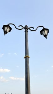 A vintage-style street lamp post with two lanterns on curved arms, set against a clear blue sky with a few clouds.