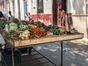 Street stall with various vegetables and fruits, houses and people in the background of a busy alley.