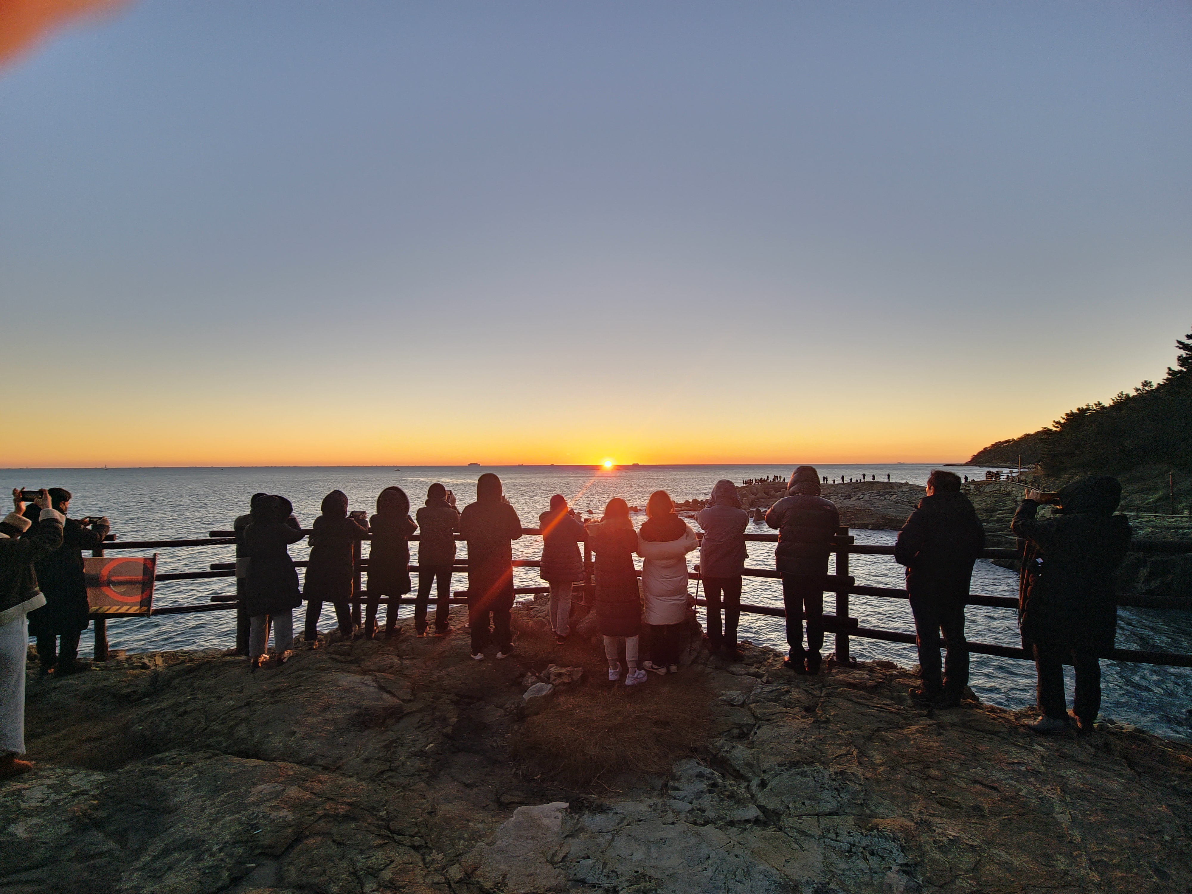 A group of people stands on a rocky shore, observing a sunrise over the ocean. The sky is clear, and the sun is just above the horizon, casting a warm glow. Igidae sunrise, Busan, South Korea