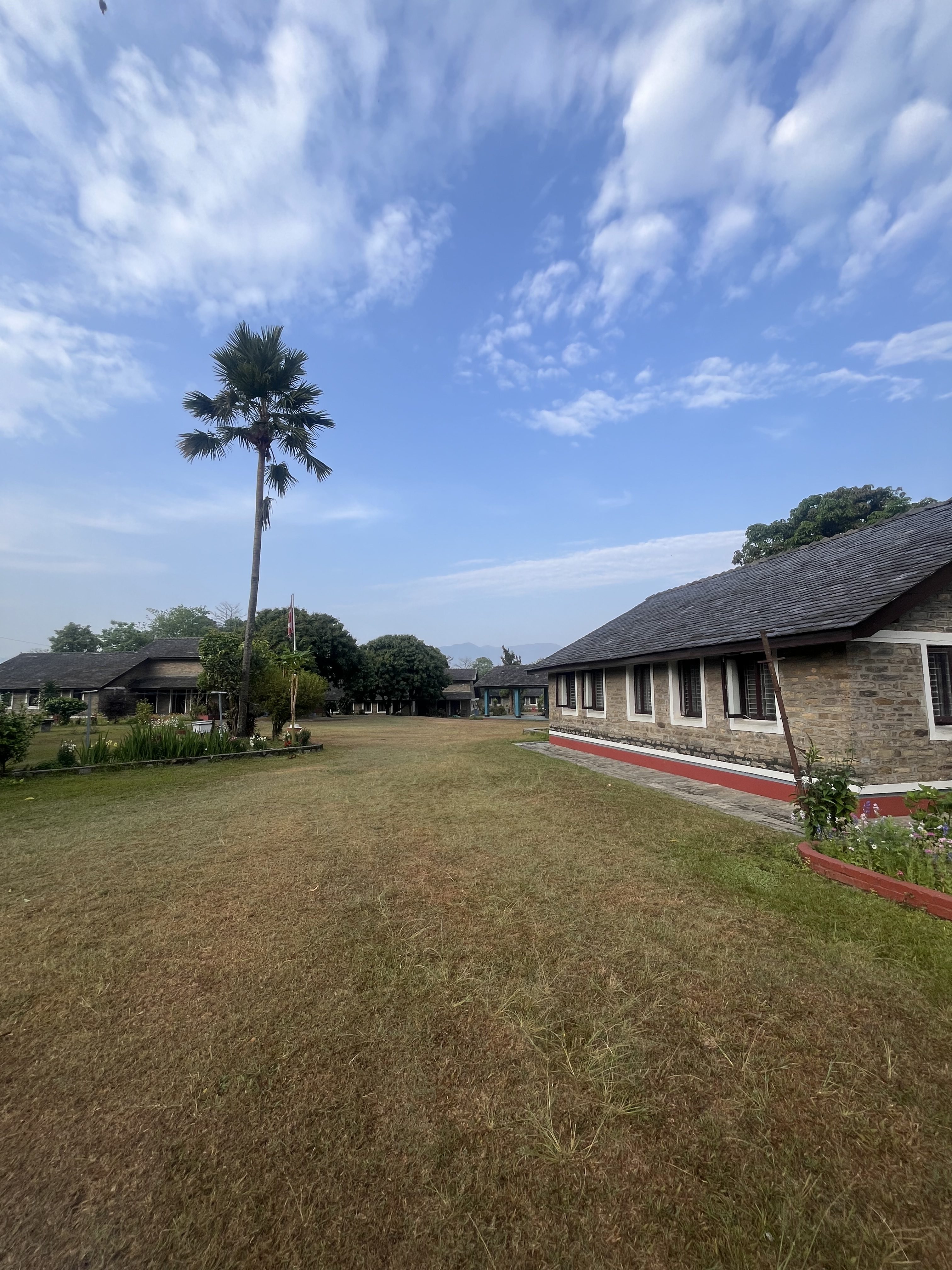 A scenic view of a grassy lawn leading to several stone buildings under a partly cloudy blue sky. A tall palm tree stands on the left, and there are gardens with small plants and flowers near the buildings.