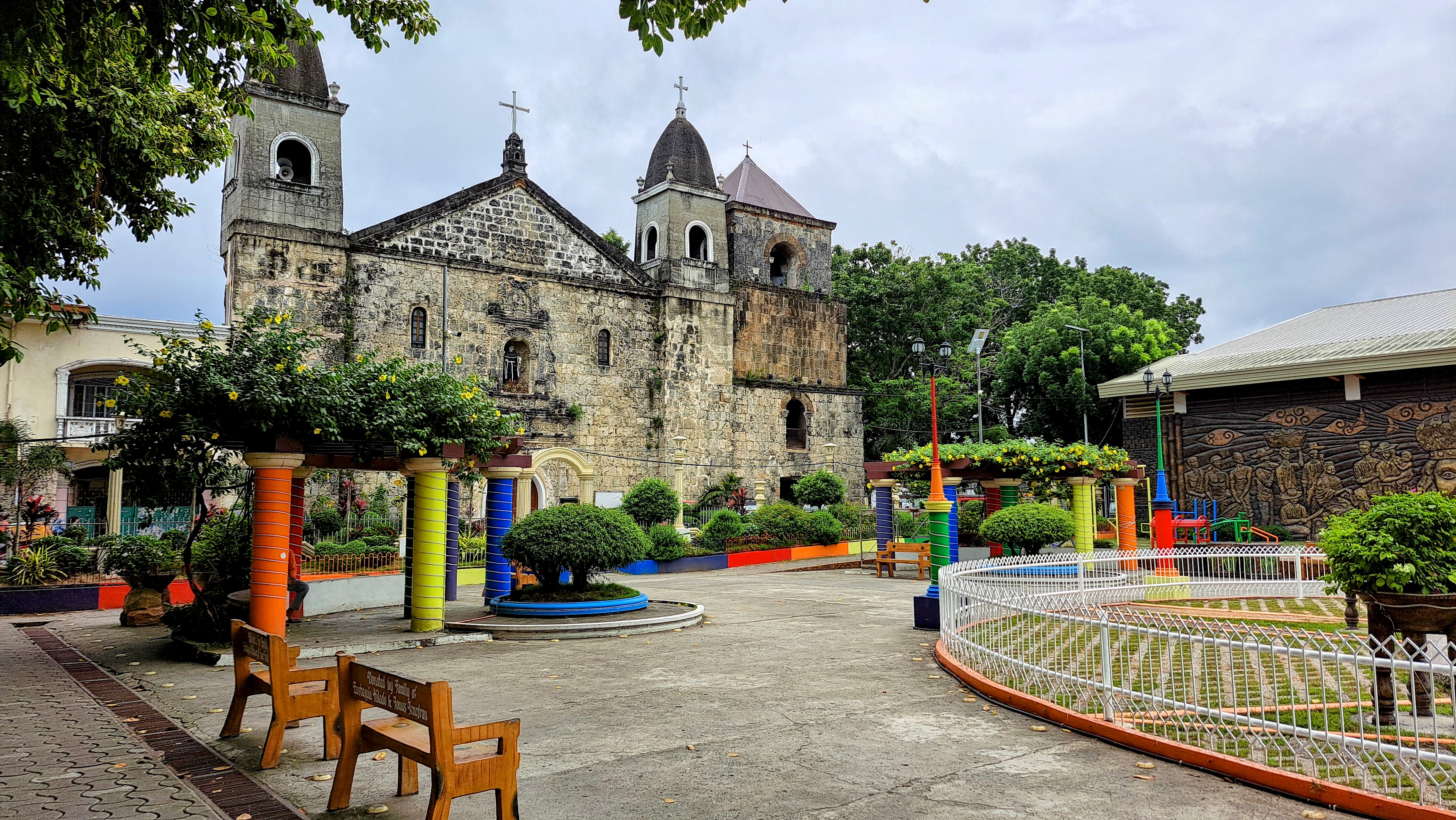 Historic stone church with two bell towers, surrounded by vibrant greenery and colorful columns in an open courtyard setting. Benches and plants decorate the foreground, with a paved path leading to the church entrance.