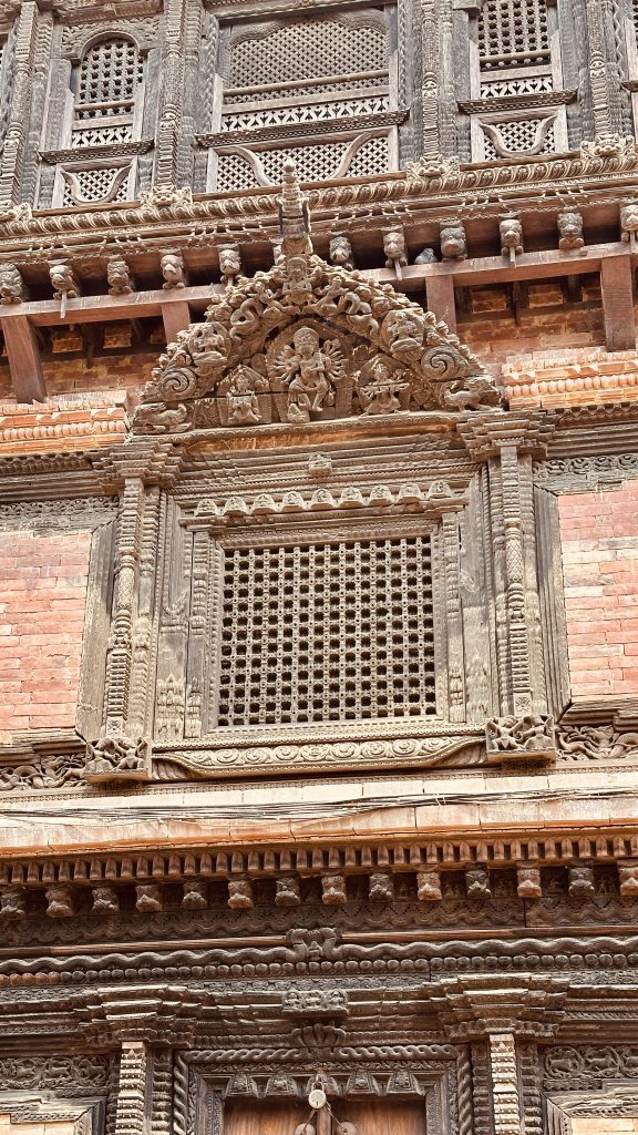 A close-up view of Bhaktapur museum intricately carved wooden architecture featuring a decorative window with a lattice design. Above the window, there are detailed carvings of figures and animals, framed by ornate borders. The surrounding walls display additional carvings and textures, showcasing traditional craftsmanship