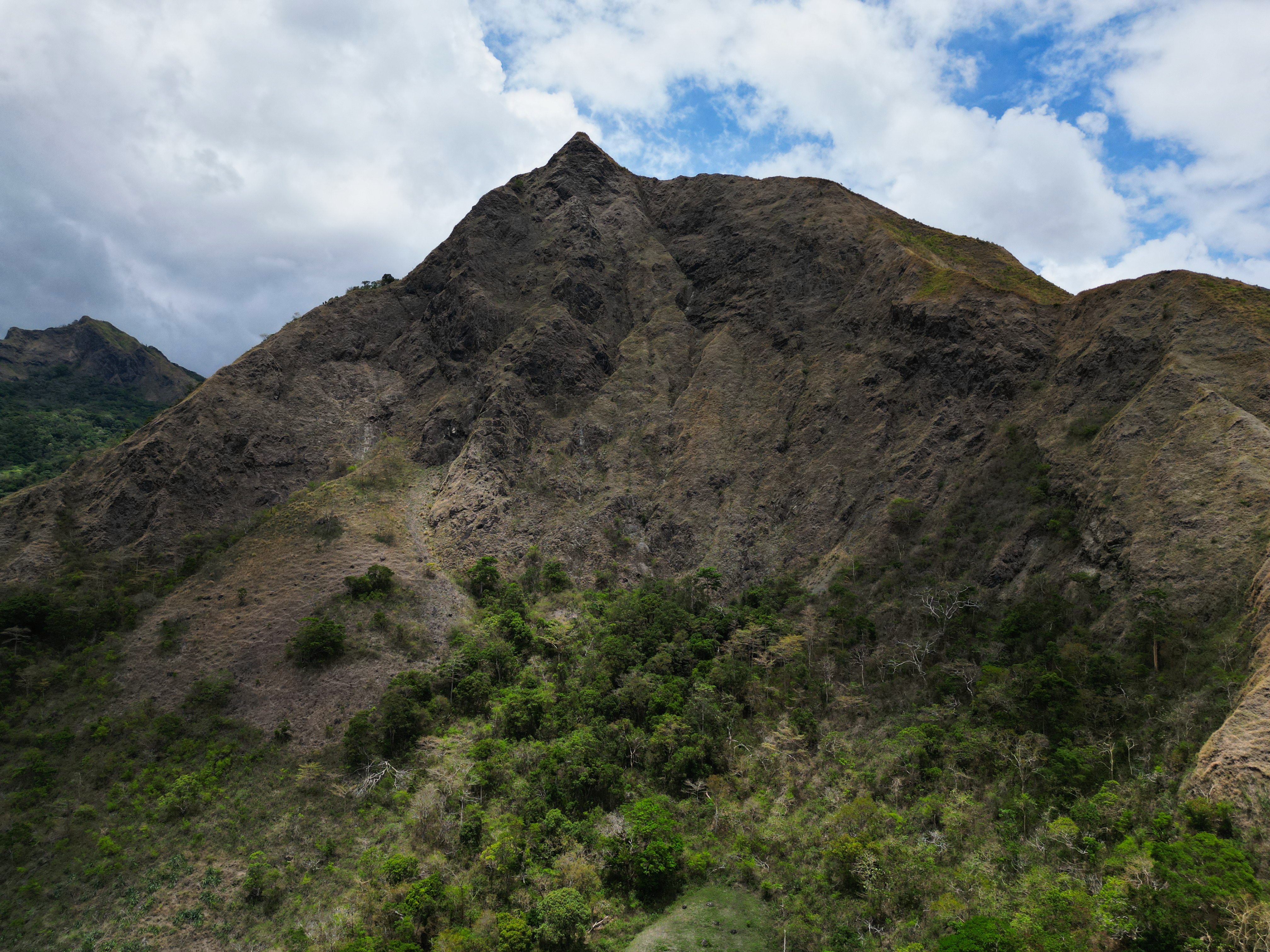 A steep, rocky mountain peak surrounded by lush greenery is set against a partly cloudy sky. The lower slopes are covered with dense vegetation, creating a contrast with the rugged rocks above.