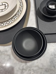 A selection of dinnerware displayed on a table. In the foreground, three black stacking bowls are nested together.