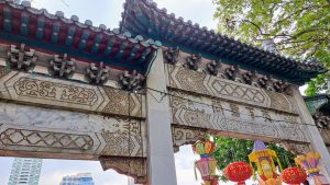 A decorative archway featuring intricate carvings and ornate designs is shown, with a colorful roof adorned with traditional patterns. Below the arch, vibrant paper lanterns in various shapes and colors hang, surrounded by lush greenery.