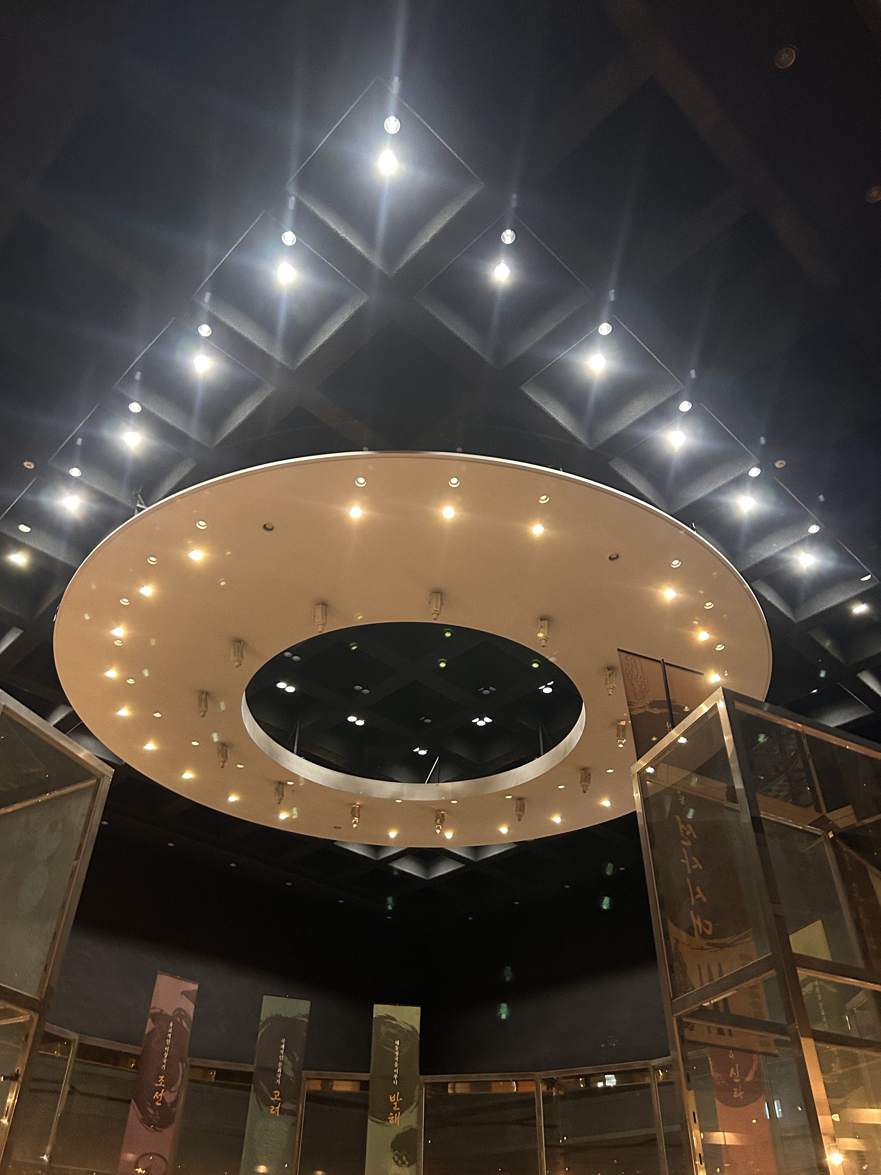 The image shows a modern architectural interior with a circular ceiling feature illuminated by bright lights. The ceiling has a grid pattern, with an elongated circular opening in the center. Below, glass panels create a transparent enclosure, and colorful banners hang from the walls, displaying stylized text in various hues.