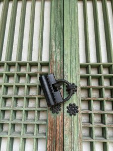 A close-up view of a weathered wooden door featuring vertical green slats and a grid pattern on one half. The door is adorned with a black metal latch and three decorative flower-shaped metal pieces near the latch, contrasting against the green wood and light-colored wall behind.