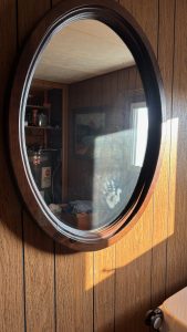 An oval mirror on a wall, the sun making a square of light in the lower right, with a child's handprint visible in the mirror.