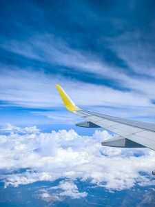 View from an airplane window showing a portion of the aircraft's wing with a yellow tip, flying above a landscape covered in fluffy white clouds and a vibrant blue sky.