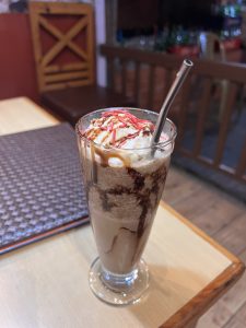 A tall glass of iced chocolate drink with whipped cream, chocolate syrup, and red sprinkles on top, accompanied by a metal straw, placed on a wooden table with a chair in the background
